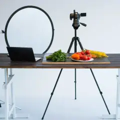 A tabletop photography setup with a camera on a tripod, a laptop, and fresh ingredients like tomatoes and pasta on a wooden table.