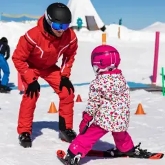 Maestro di sci insegna a bambini a sciare su pista innevata, con coni colorati e bandierine sullo sfondo.