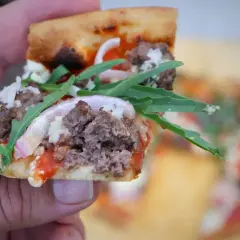 Kawałek pysznej pizzy 30 cm z mielonym mięsem, rukolą i czerwoną cebulą, trzymany w dłoni.