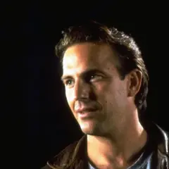 Kevin Costner jung, mit nachdenklichem Blick und brauner Lederjacke vor dunklem Hintergrund.