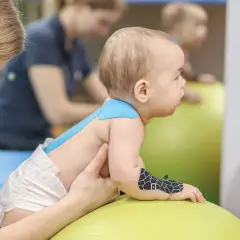 Dziecko na piłce rehabilitacyjnej, z taśmami kinesiotaping. Terapeuta wspiera malucha podczas ćwiczeń.