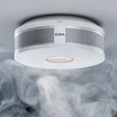Ein Gira Rauchmelder mit orange blinkendem Ring, umgeben von aufsteigendem Rauch.