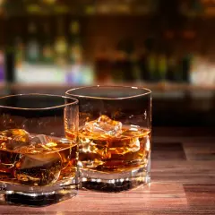 Dwa szklanki whisky z lodem na drewnianym blacie. W tle rozmyty bar, sugerujący atmosferę, która napędza przyczyny popularności whisky rzemieślniczej.