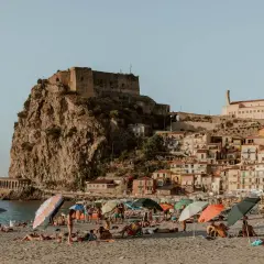 A sunny beach with people relaxing under umbrellas, overlooking a cliffside town with a castle. This view is a perfect destination for those wondering how to get to Calabria, Italy.