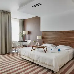 Przytulny pokój hotelowy z dwoma łóżkami, stolikiem i fotelami, ozdobiony drewnianym zagłówkiem i pasiastym dywanem.