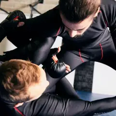 Dwóch zawodników w czarnych rashguardach walczy na macie. Jeden próbuje obalić drugiego, który broni się nogami. To zacięta walka w kategorii waga piórkowa.