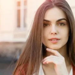 Portret młodej kobiety z dłonią pod brodą, w tle budynek.