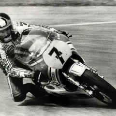 Wyścigowy motocykl Suzuki z numerem 7 pochylony w zakręcie, z zawodnikiem w kasku. To fragment historii motocykla.