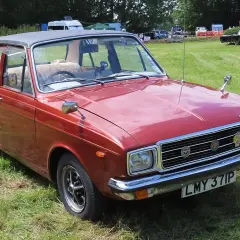 Röd Hillman bil med svart vinyltak, parkerad på en gräsmatta vid ett evenemang.
