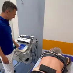 Fizjoterapeuta ustawia parametry na aparacie do elektroterapii, co to jest zabieg prądami. Pacjent leży na kozetce.