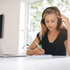 Mujer con auriculares escribe en un cuaderno, con luz natural entrando por la ventana. La luz suave es ideal, preguntándose cuál es la mejor luz para estudiar.