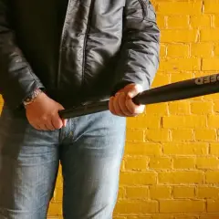 Mężczyzna w czarnej kurtce i jeansach trzyma kij baseballowy z napisem "SPEED". Tło to żółta ceglana ściana.