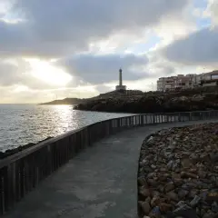 Camino de piedra y madera bordeando el mar, con un faro al fondo. La carretera subida al faro invita a pasear al atardecer.