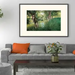 A modern impressionism painting of a serene riverbank with lush green grass and trees, framed above a grey sofa with orange and grey cushions.