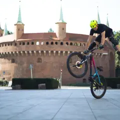 Wykonuje trik na rowerze po krakowsku, z zabytkowym Barbakanem w tle.
