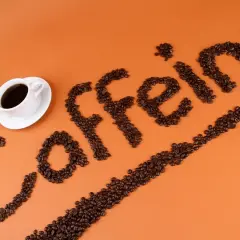Ziarna kawy ułożone w napis "caffeine" obok filiżanki kawy. Zastanawiasz się, ile kofeiny ma łyżeczka kawy mielonej?