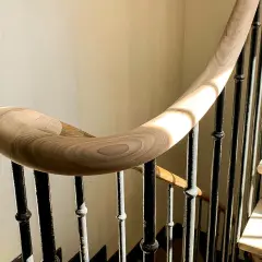Courbe élégante d'une main courante en bois sur une volée d'escalier aux barreaux de fer forgé, baignée de lumière.