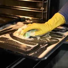 Main tenant avec des gants jaunes, nettoie une plaque de cuisson savonneuse pour enlever odeur four.