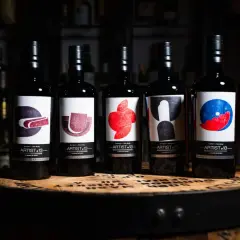 Cinq bouteilles de blend whisky Artist-13, ornées d'étiquettes artistiques colorées, reposent sur un fût de whisky.