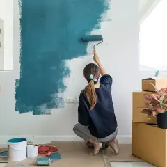 Mujer pintando una pared de azul, rodeada de cajas. Es la mejor época para pintar la casa y darle un nuevo aire.