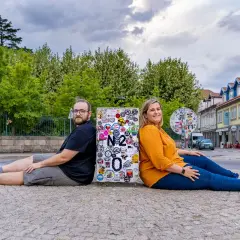 Pareja sentada de espaldas en un poste con pegatinas, celebrando su viaje por la Estrada Nacional 2 en Portugal.