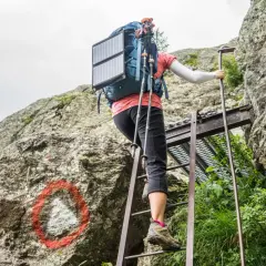 A hiker ascends metal ladders on the Tour du Mont Blanc, using trekking poles and a solar panel on their backpack.