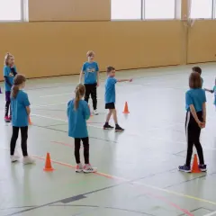 Kinder in blauen T-Shirts üben in der Turnhalle, während ein Trainer Anweisungen gibt. Tip tap, die Füße bewegen sich im Takt.