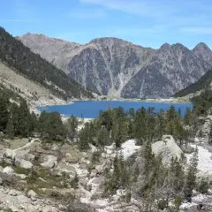 Lago azul rodeado de montañas y pinos, con un pequeño refugio en la orilla. El refugio Oulettes de Gaube es un destino popular para excursionistas.