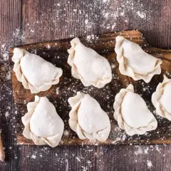 Surowe pierogi na desce, gotowe do przechowywania w lodówce. Obok łyżka z mąką.