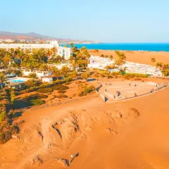 Vacker vy över Dunas de Maspalomas med vita hotell, palmer och gyllene sanddyner mot det blå havet.