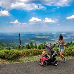 Kobieta pcha wózek z dzieckiem po górskiej ścieżce z widokiem na dolinę.