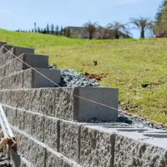 Budowa muru oporowego z bloczków betonowych na zboczu. Widać drabinę i sznur budowlany.