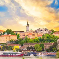 Widok na duże miasto w Serbii z rzeką i statkami. W tle widać kościół z zieloną kopułą i wieżą.
