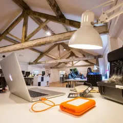 Biuro z drewnianymi belkami na suficie. Praca w pomieszczeniu bez dostępu światła dziennego, oświetlonym lampami. Na biurku laptop, dysk zewnętrzny i sprzęt do nagrywania.