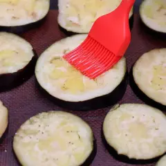 Rodajas de berenjenas listas para ser horneadas, untadas con aceite y especias con un pincel rojo.