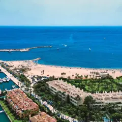 Widok na marinę pełną jachtów i piaszczystą plażę, gdzie rozciąga się złoty wybrzeże. W tle błękitne morze i horyzont.