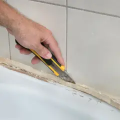 Ręka z nożem usuwa stary silikon z wanny. Dowiedz się, czym usunąć silikon z wanny, by łazienka znów wyglądała jak nowa.