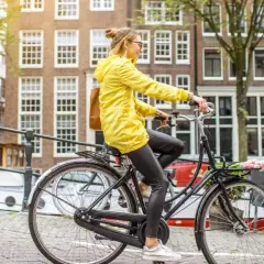 Vrouw in gele jas rijdt op een simpele stadsfiets door Amsterdam, met bloemen in het mandje.