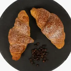 Dos cruasanes dorados espolvoreados con azúcar glas, junto a granos de café. Una delicia para aprender como hacer cruasanes.