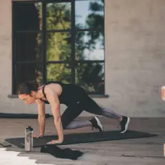 Femme en position de planche, genou vers poitrine, effectuant un exercice polyarticulaire. Bouteille d'eau et téléphone à proximité.