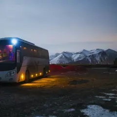 Autobus z zapalonymi światłami na tle ośnieżonych gór o zmierzchu.