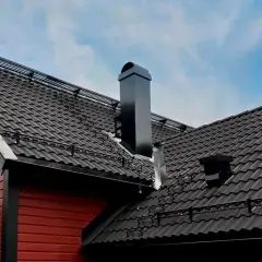 Svart, modern skorsten på ett tak med mörka takpannor. En röd träfasad syns till vänster.
