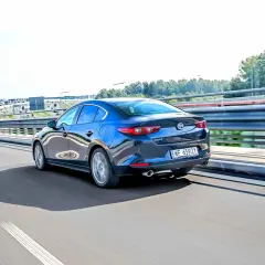 Ciemnoniebieska Mazda 3 sedan jedzie po autostradzie, odbijając światła i otoczenie.