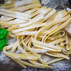 Świeży, domowy makaron bez glutenu, posypany mąką, z listkami bazylii i drewnianą łyżką na drewnianym blacie.