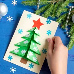 Dziecko tworzy kartkę z papieru, pokazując, jak zrobić ozdoby świąteczne z papieru. Zielona choinka z płatkami śniegu i czerwoną gwiazdą.