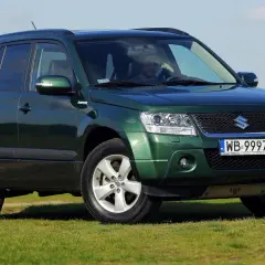 Zielony Suzuki Grand Vitara na tle łąki i błękitnego nieba. Kierowca za kierownicą.