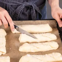 Hände bereiten Teiglinge für Laugenstangen zum Aufbacken vor. Ein Messer schneidet in einen Teigling, grobes Salz liegt bereit.