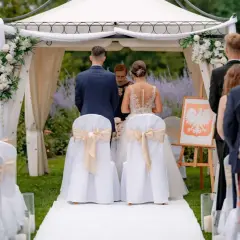 Uroczysta ceremonia zaślubin pod baldachimem, z gośćmi w tle. Staranna organizacja wesela widoczna w dekoracjach i przygotowaniu miejsca.