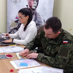 Żołnierz w mundurze pieczętuje dokumenty, podczas gdy kobieta pracuje przy laptopie. To część procesu kwalifikacji wojskowej.
