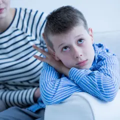 Chłopiec z autyzmem opiera głowę na ramieniu, patrząc w górę. Obok siedzi kobieta. Obraz symbolizuje poszukiwanie przyczyn autyzmu u dzieci.